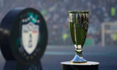 Shiny silver and green trophy on a pedestal with a blurred stadium crowd in the background, suggesting a championship award.