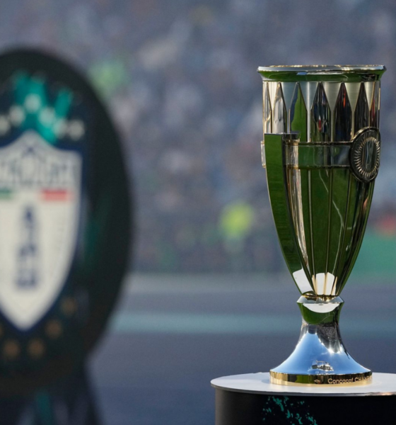 Shiny silver and green trophy on a pedestal with a blurred stadium crowd in the background, suggesting a championship award.