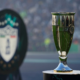 Shiny silver and green trophy on a pedestal with a blurred stadium crowd in the background, suggesting a championship award.