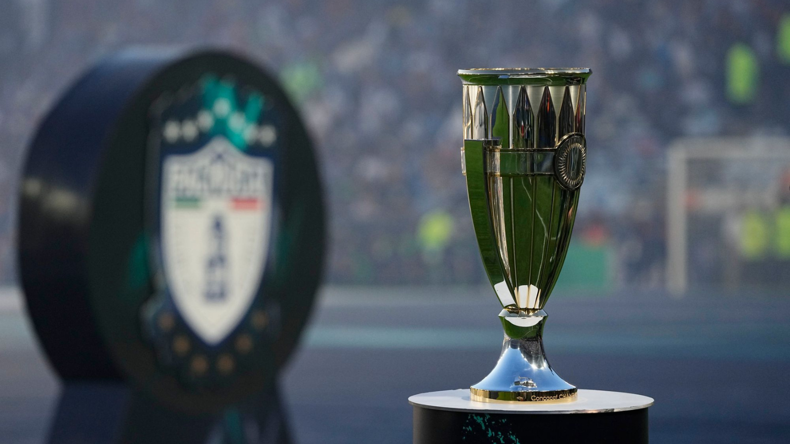 Shiny silver and green trophy on a pedestal with a blurred stadium crowd in the background, suggesting a championship award.