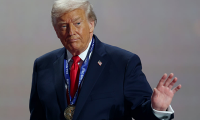 Older man in a navy suit and red tie wears a large medal on a blue lanyard at a ceremony, raising his right hand.