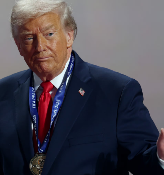 Older man in a navy suit and red tie wears a large medal on a blue lanyard at a ceremony, raising his right hand.