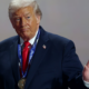 Older man in a navy suit and red tie wears a large medal on a blue lanyard at a ceremony, raising his right hand.