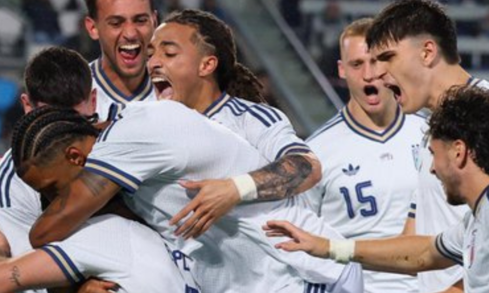 Soccer players in white uniforms celebrate with a group hug on the field, arms around each other in joy.