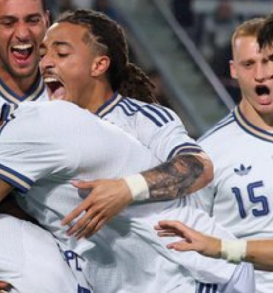 Soccer players in white uniforms celebrate with a group hug on the field, arms around each other in joy.