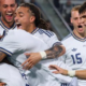 Soccer players in white uniforms celebrate with a group hug on the field, arms around each other in joy.