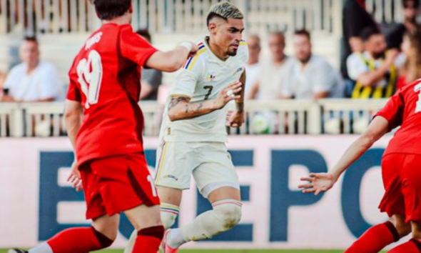 Soccer player in a white kit runs with the ball while two players in red chase on a sunny field with spectators in the background.