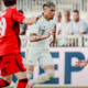 Soccer player in a white kit runs with the ball while two players in red chase on a sunny field with spectators in the background.