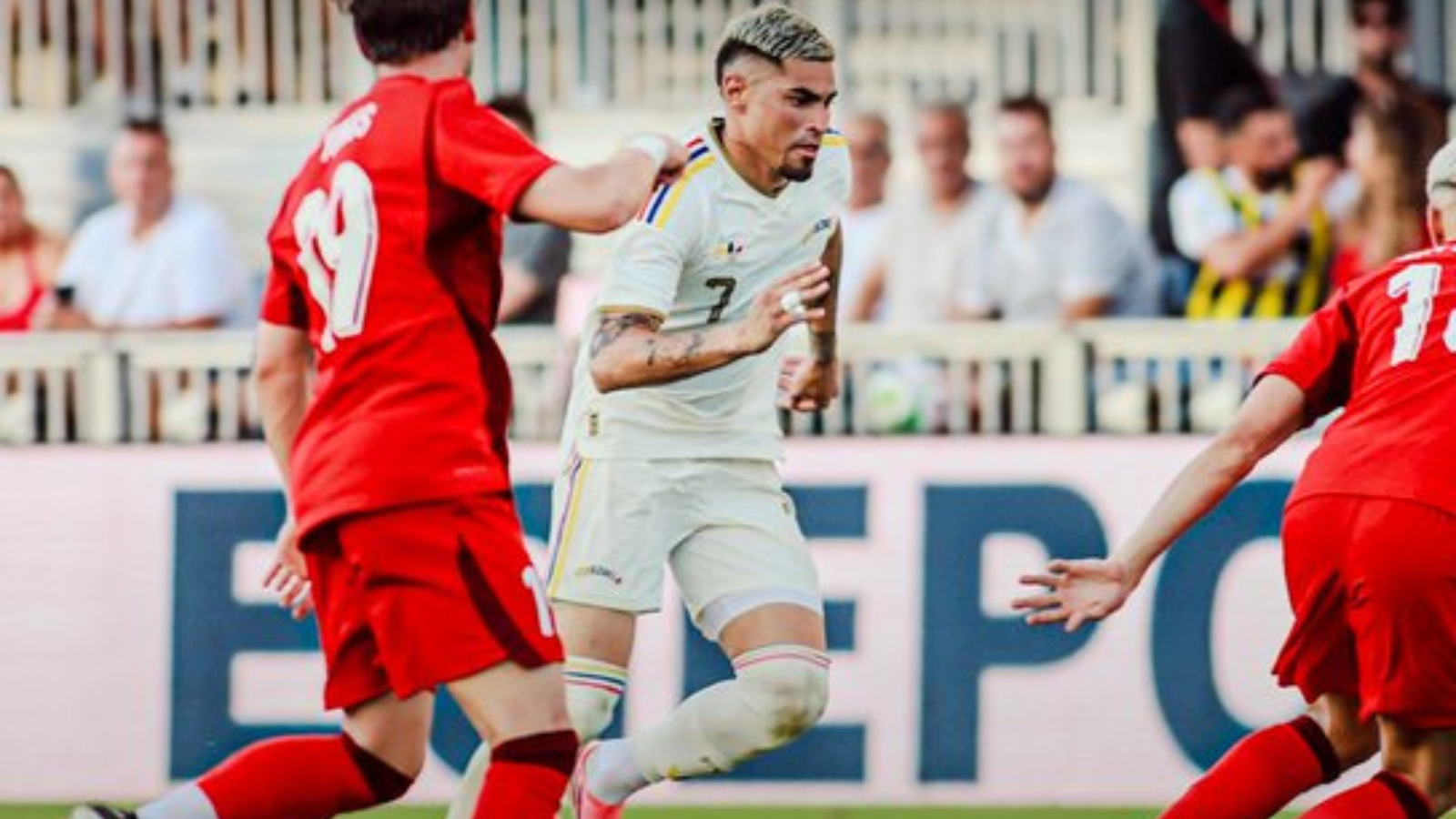 Soccer player in a white kit runs with the ball while two players in red chase on a sunny field with spectators in the background.