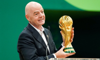 Bald man in a dark suit smiles while presenting a gold World Cup trophy at a podium with a microphone nearby.