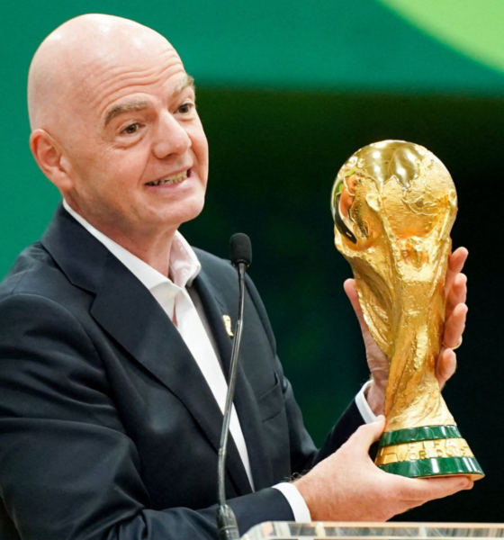Bald man in a dark suit smiles while presenting a gold World Cup trophy at a podium with a microphone nearby.