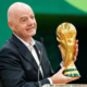 Bald man in a dark suit smiles while presenting a gold World Cup trophy at a podium with a microphone nearby.
