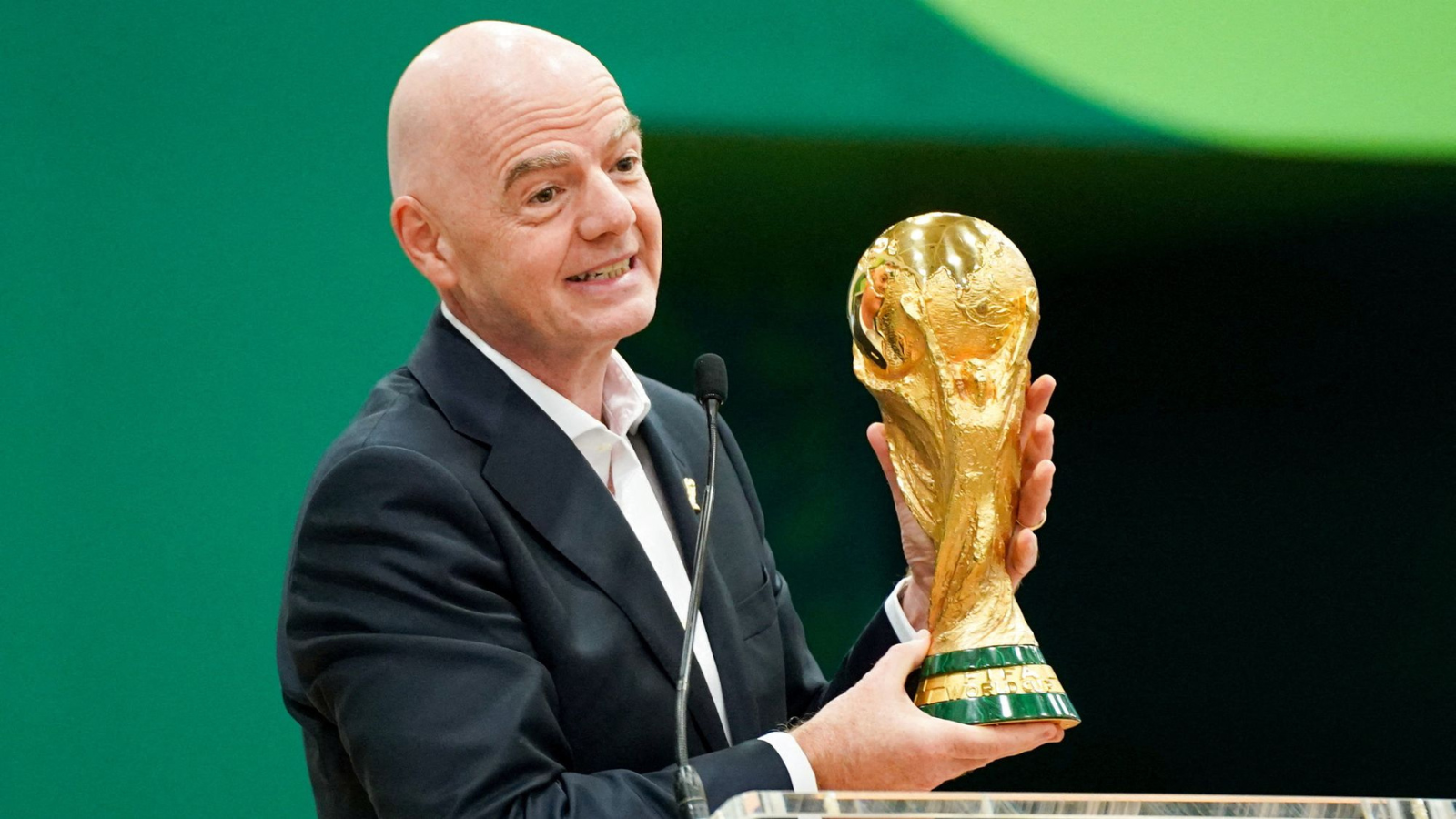 Bald man in a dark suit smiles while presenting a gold World Cup trophy at a podium with a microphone nearby.
