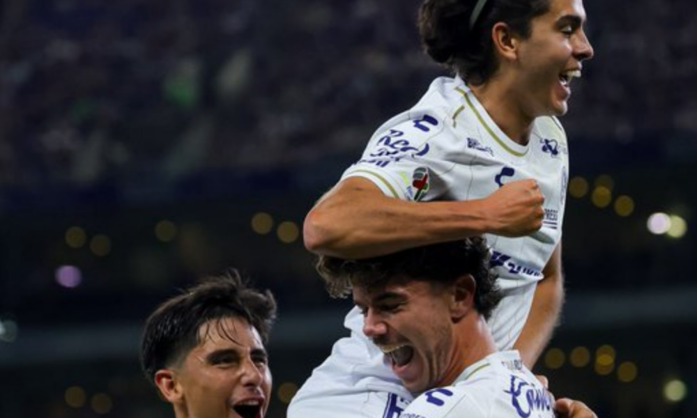 Three soccer players in white jerseys celebrate a goal, one on another's shoulders, smiling and shouting with joy in a stadium.