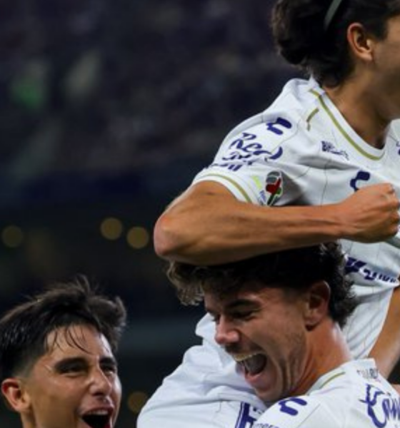 Three soccer players in white jerseys celebrate a goal, one on another's shoulders, smiling and shouting with joy in a stadium.