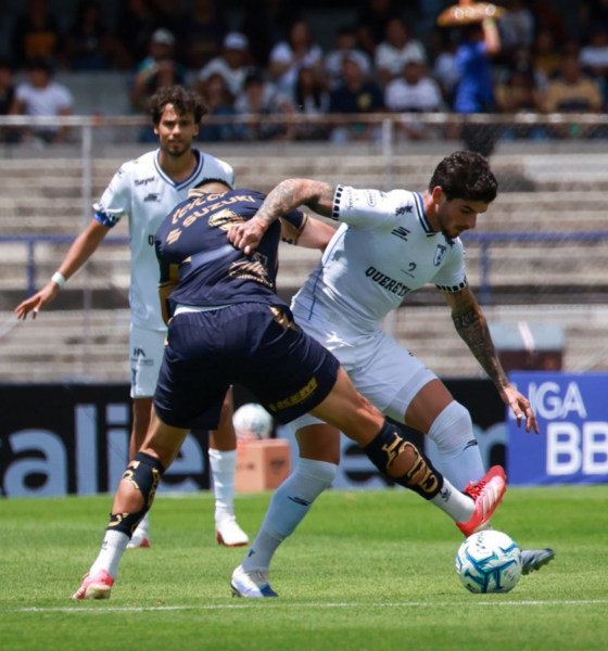 Two soccer players duel for the ball on a sunny field, one in white kit blocking the other in dark blue as fans watch from stands behind them.