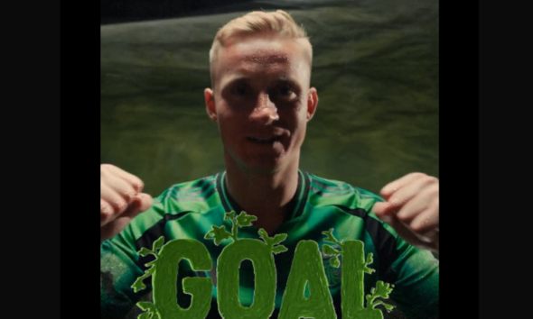 Young man in a green soccer jersey shows a large decorative sign reading GOAL in front of him, celebrating a score.